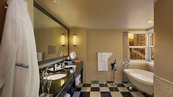 Baño de hotel con piso a cuadros, un espejo grande, dos lavamanos, una bañera y una ventana con vistas a edificios.