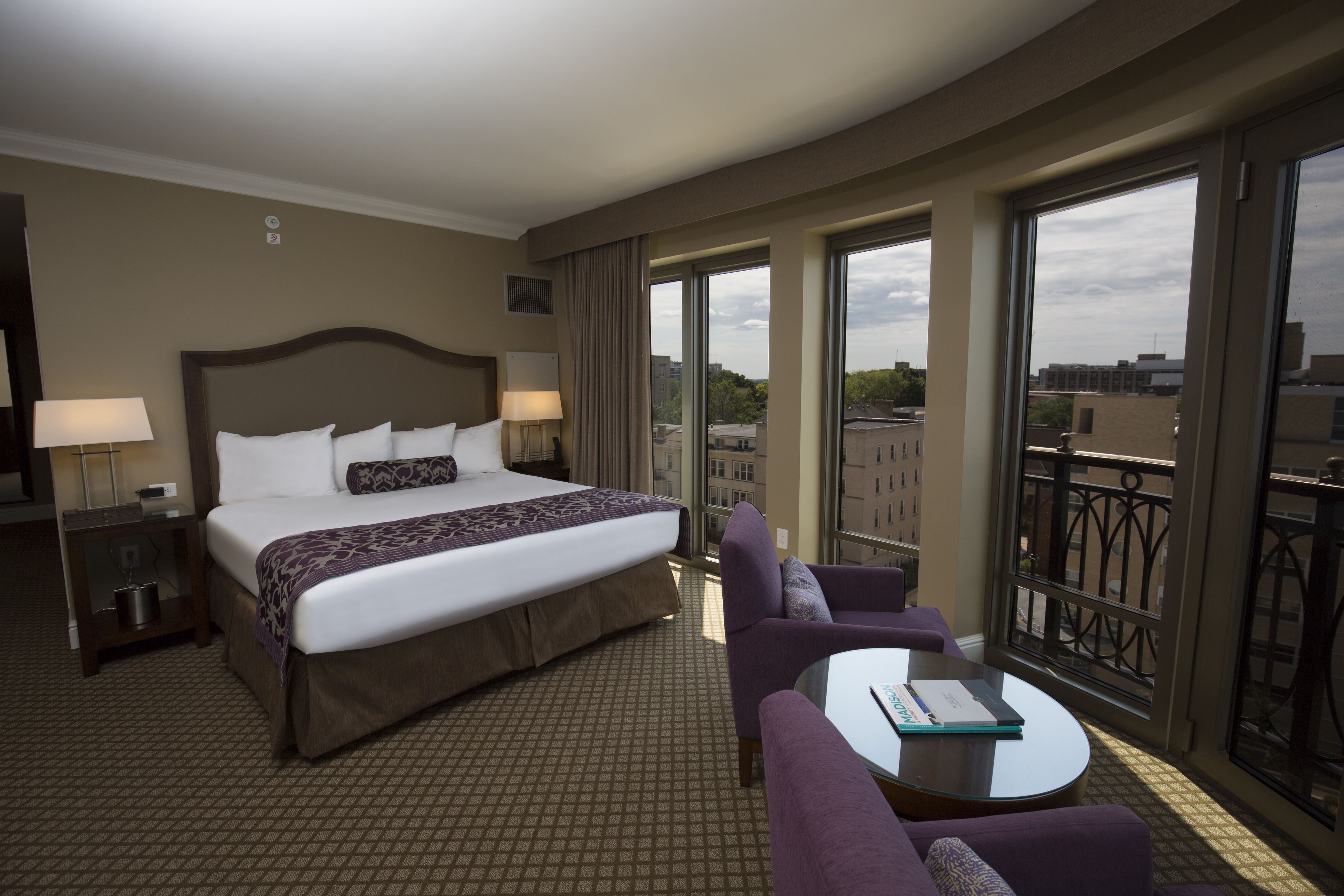 A hotel room with a large bed, a sofa, a small table, and a balcony with a view of the city.