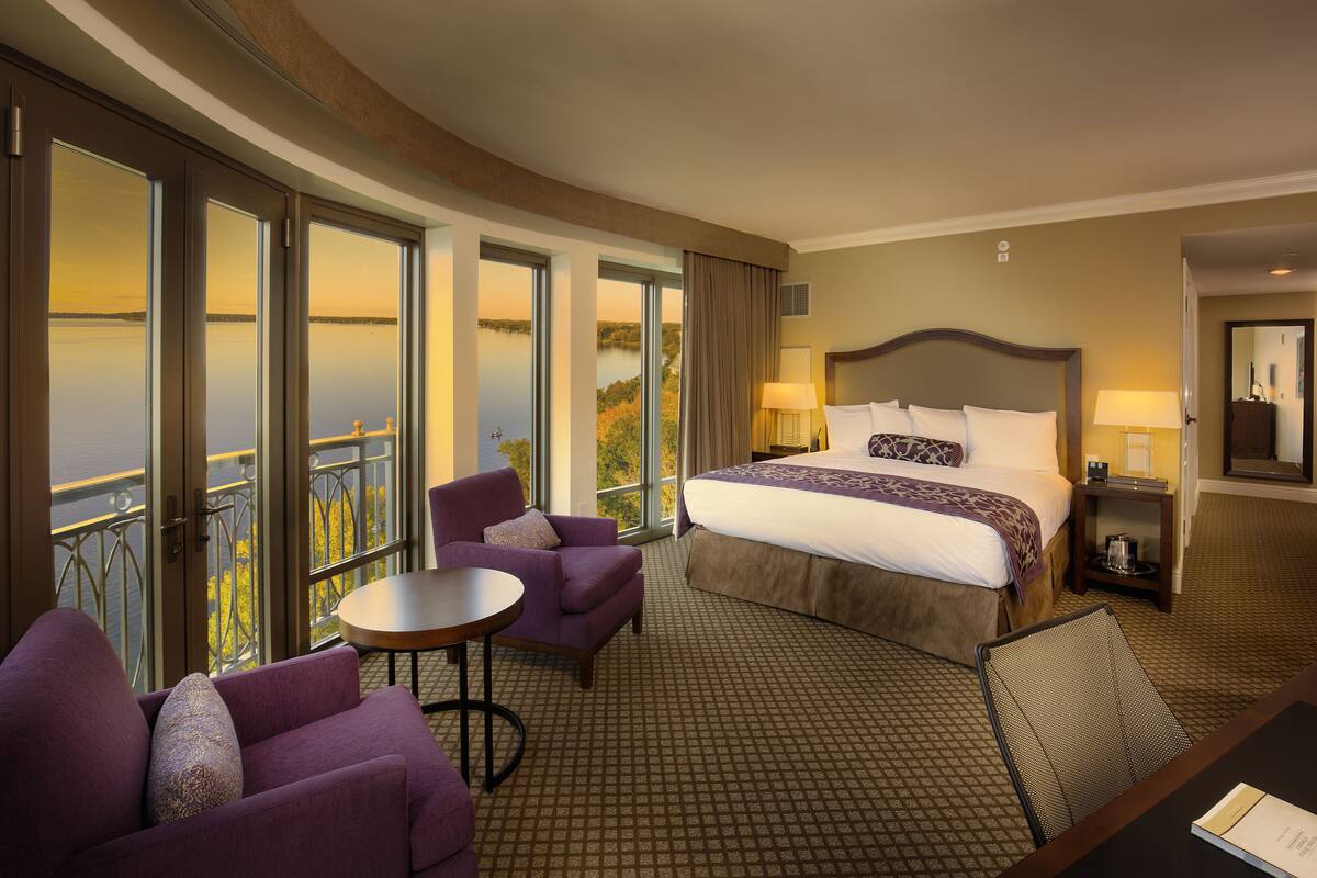 A hotel room with a large bed, a chair, a small table, and a view of a body of water through large windows.
