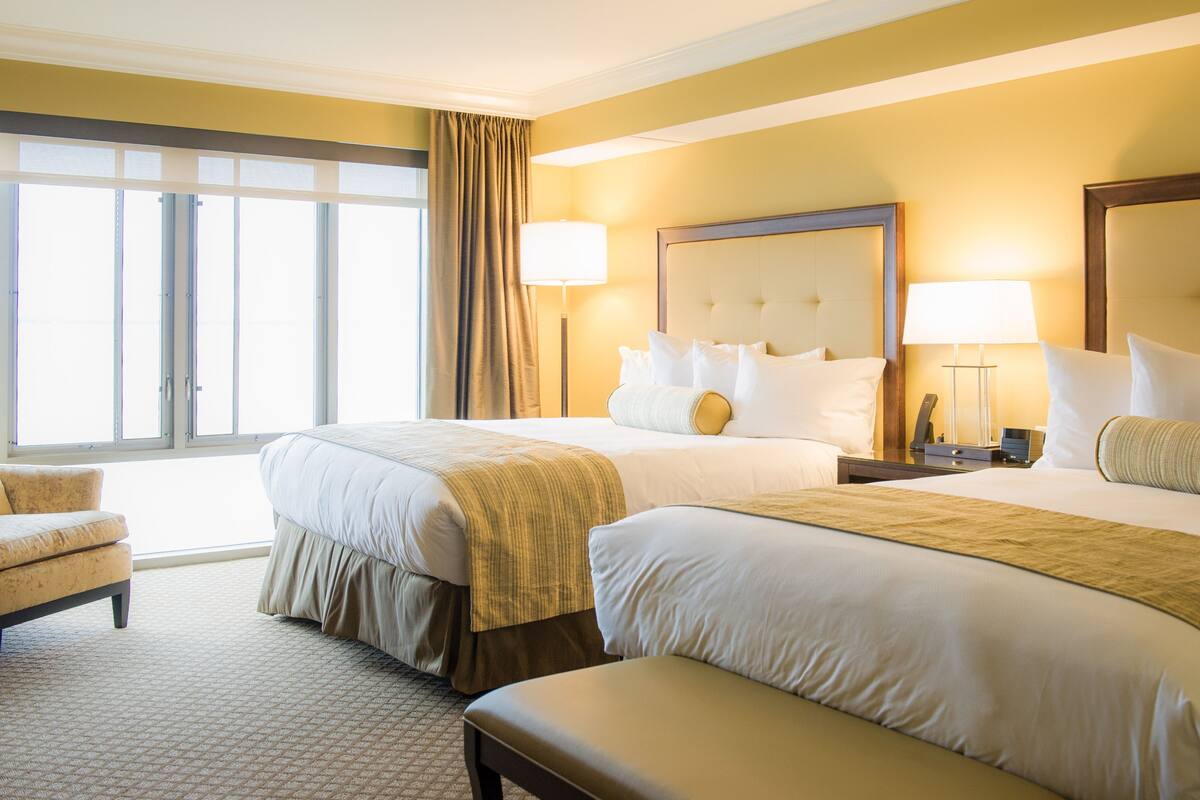 A hotel room with two beds, a large window with curtains, a chair, and a bedside table with a lamp.