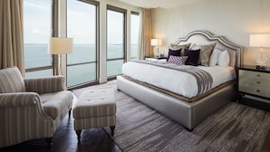A spacious bedroom with a large bed, a sitting area with a chair, and a view of the sea through large windows.