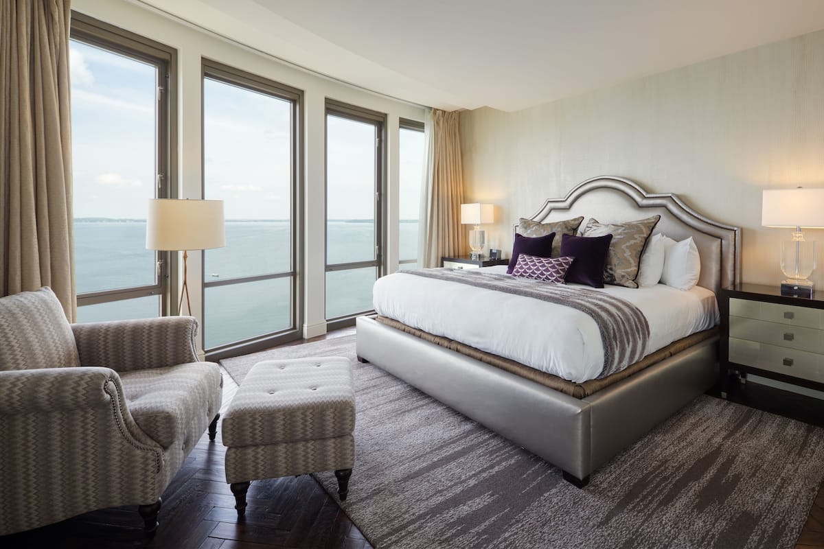 A spacious bedroom with a large bed, a sitting area with a chair, and a view of the sea through large windows.