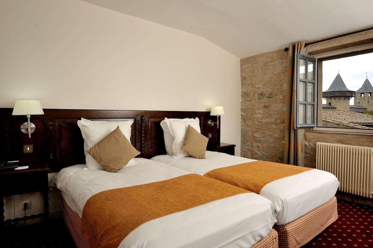 Cama doble con ropa de cama blanca y marrón, dos lámparas de noche y una ventana con vista a un castillo.