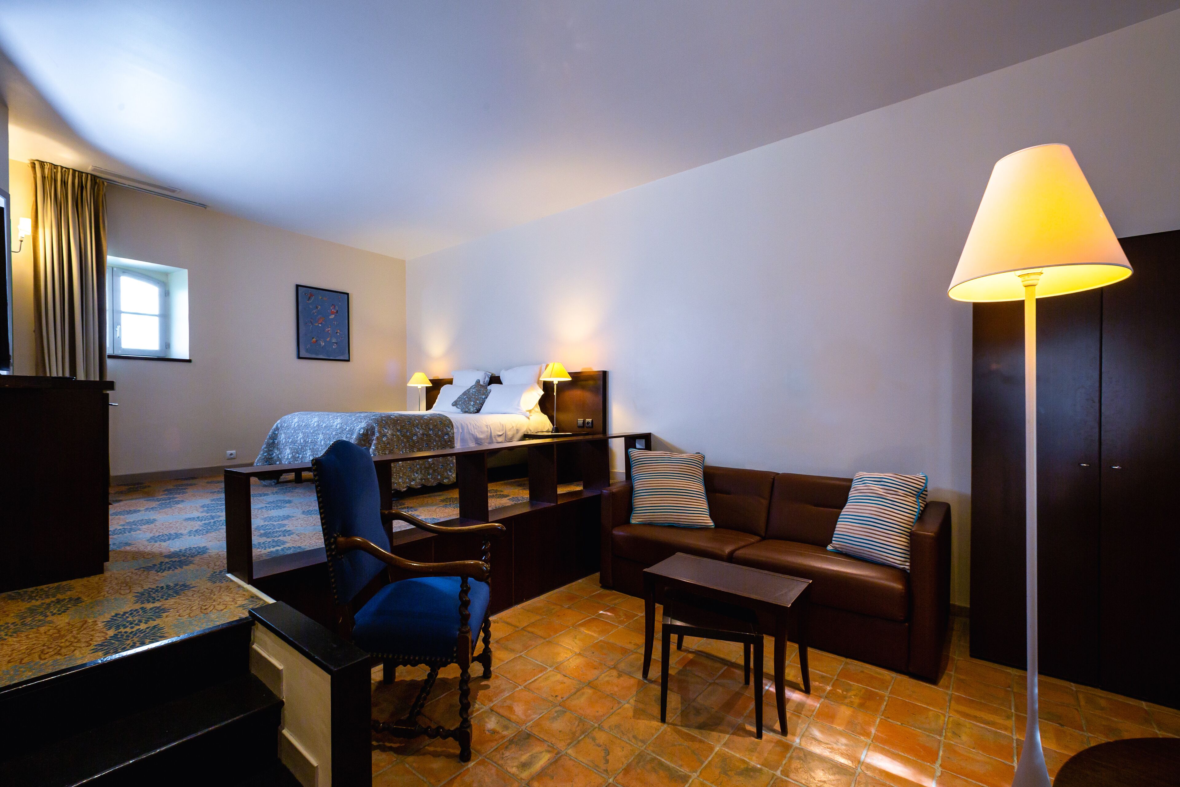 A hotel room with a bed, a sofa, a chair, a desk, and a lamp.