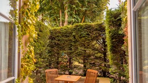 An outdoor patio with a table and chairs, surrounded by greenery and a stone floor.