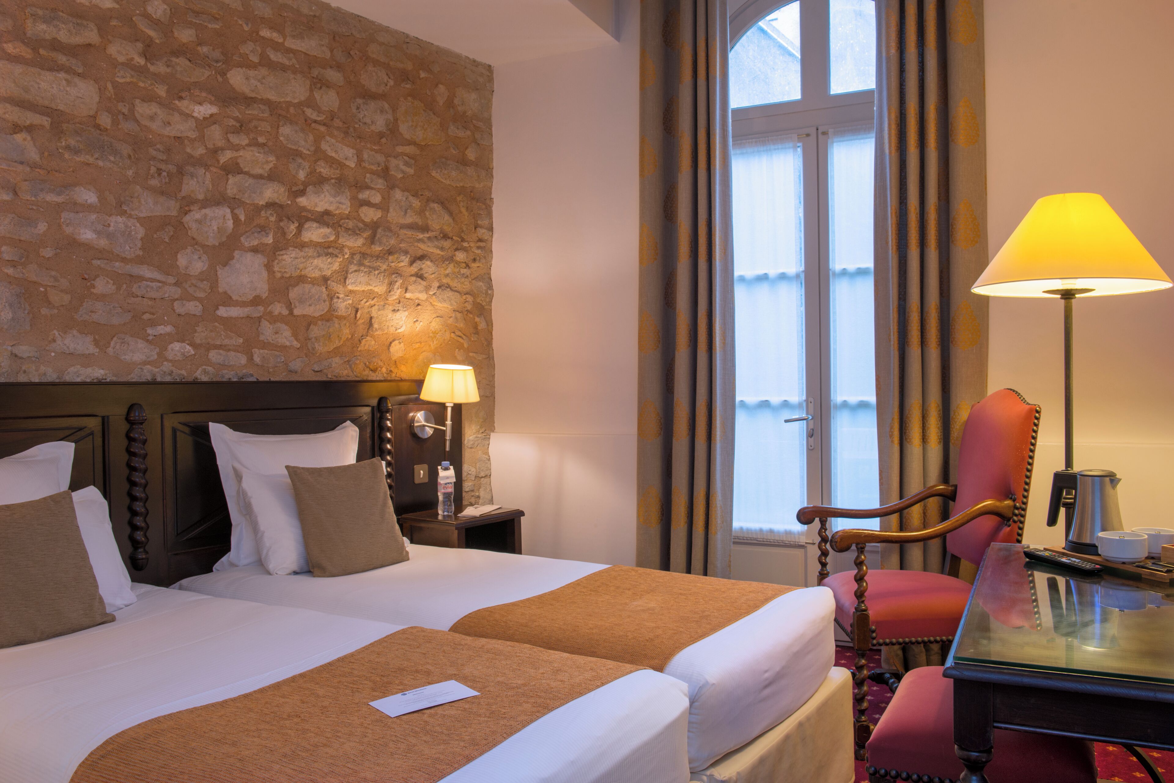 A hotel room with a stone wall, two beds with pillows, a nightstand with a lamp, a chair, a table with a kettle, and a window with curtains.