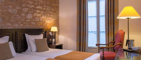 A hotel room with a stone wall, two beds with pillows, a nightstand with a lamp, a chair, a table with a kettle, and a window with curtains.