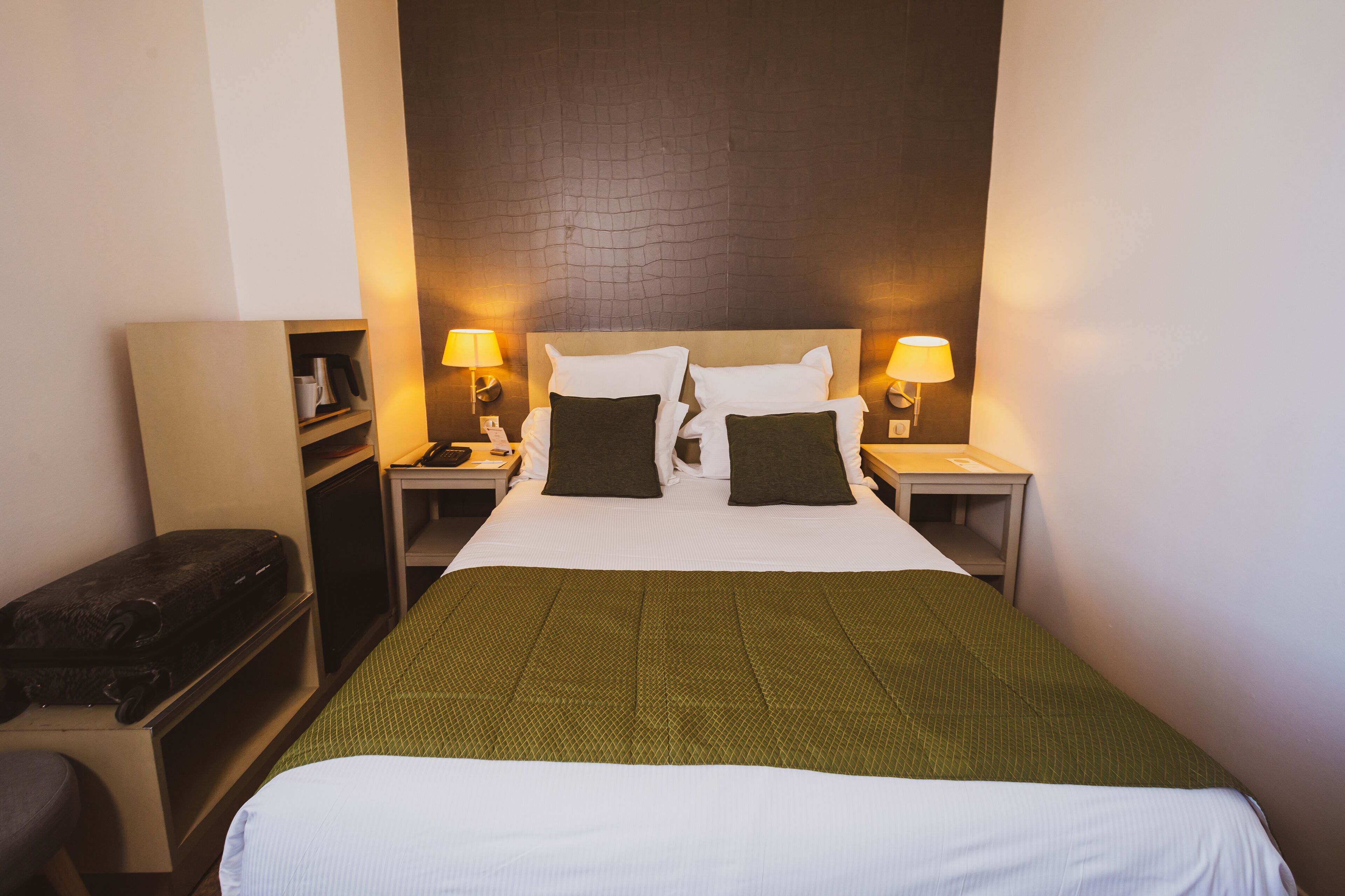 A hotel room with a large bed, two bedside tables, a nightstand, a bench, and a wall-mounted shelf.