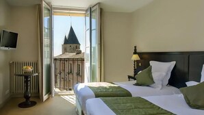 A hotel room with two beds, a table with a fruit bowl, a TV, and a view of a tower through an open door.