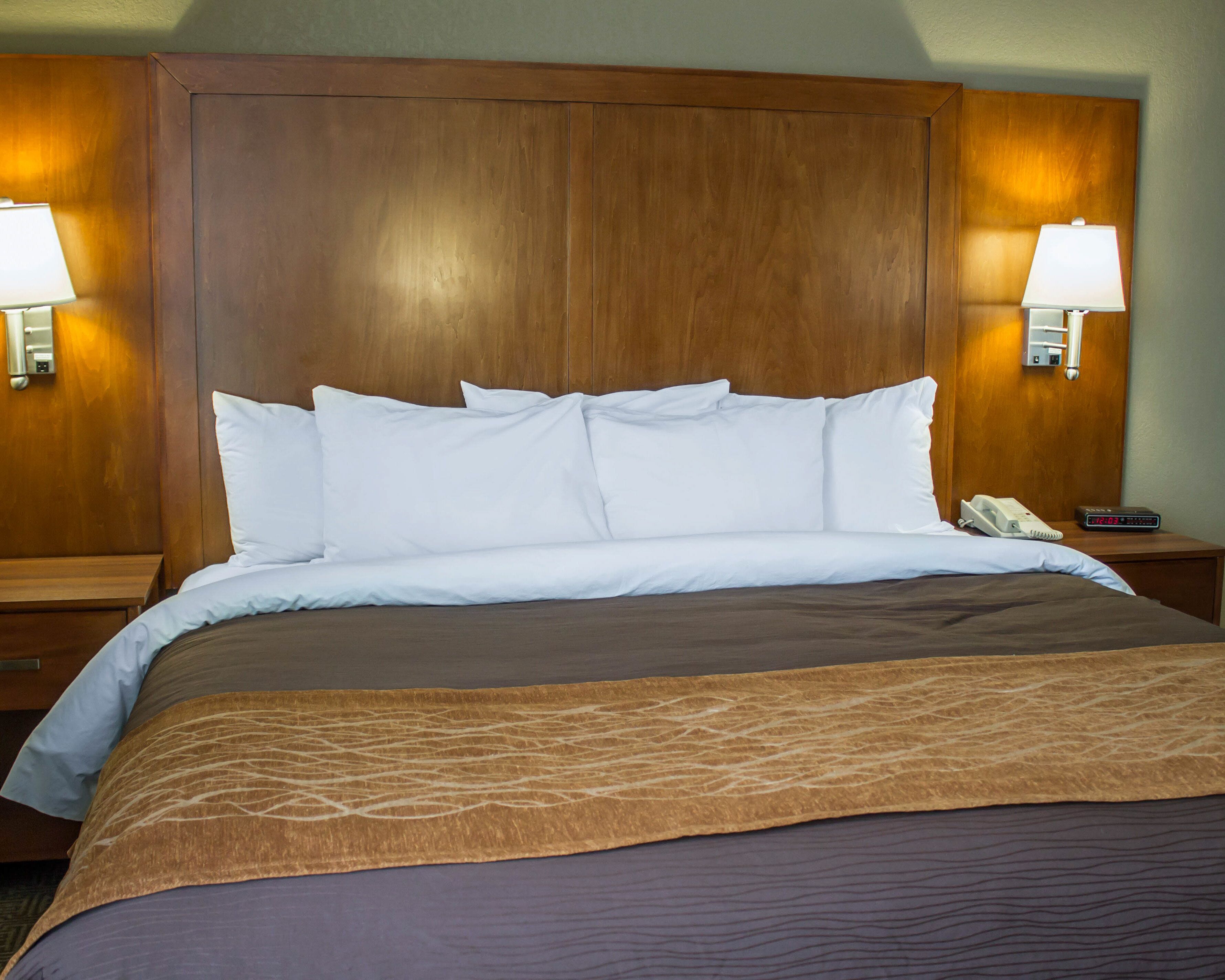 A hotel room with a large wooden headboard, white bedding, a brown throw blanket, and bedside lamps.