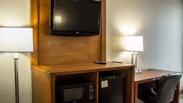 A hotel room with a wooden desk, a flat-screen TV mounted above it, a microwave, a refrigerator, and two lamps.