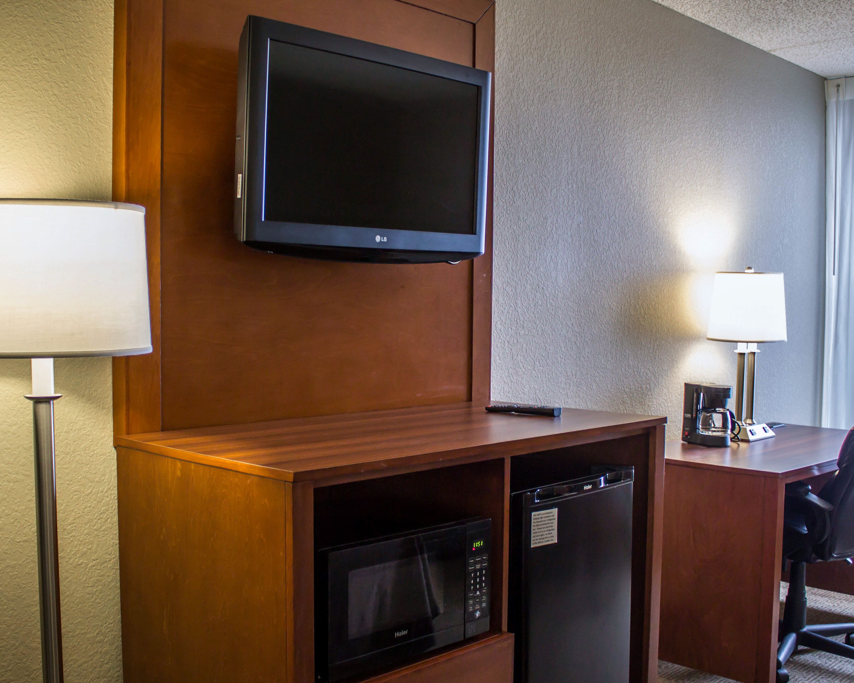 A hotel room with a flat-screen TV mounted on the wall, a microwave, a refrigerator, a desk with a lamp, and a chair.