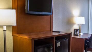 A hotel room with a flat-screen TV mounted on the wall, a microwave, a refrigerator, a desk with a lamp, and a chair.
