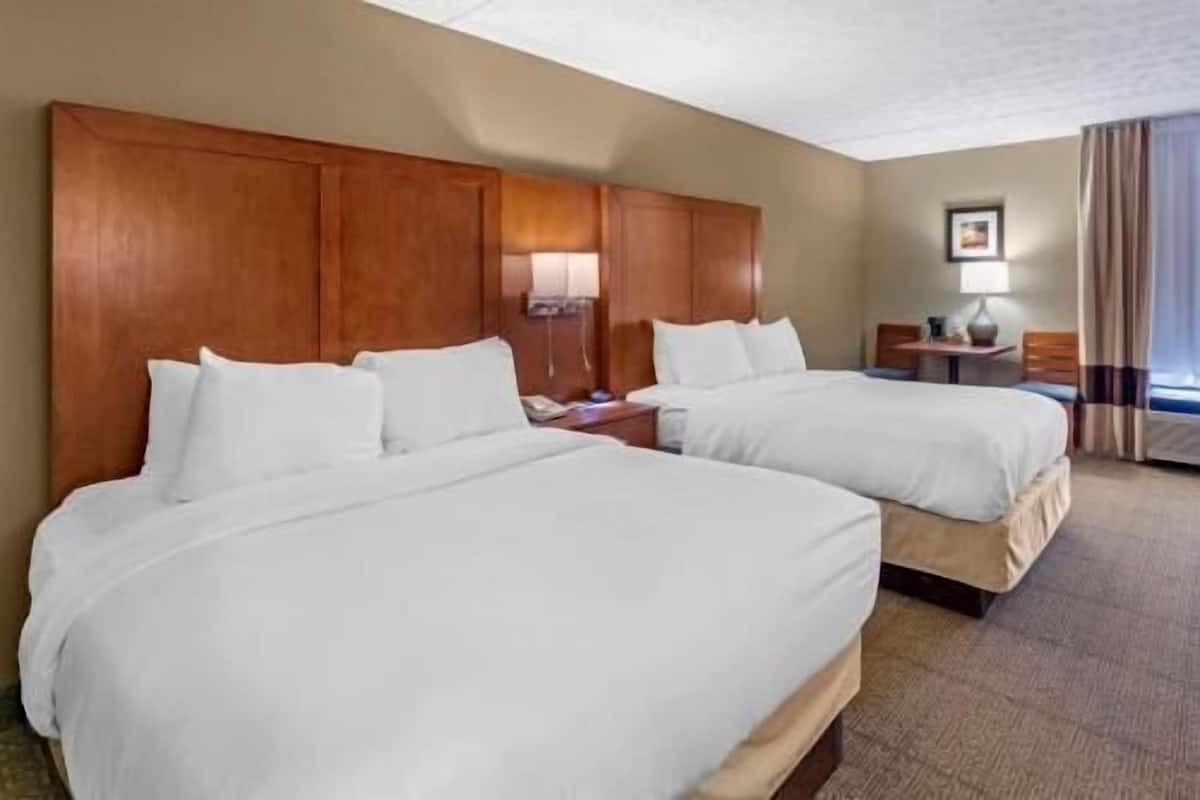 Une chambre d’hôtel avec deux lits, des têtes de lit en bois, une table de chevet avec une lampe, une petite table avec une lampe et une fenêtre avec des rideaux.