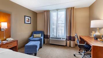 Ein Hotelzimmer mit einem Bett, einem Schreibtisch mit Lampe, einem Sessel, einer Klimaanlage und einem Fenster mit Vorhängen.