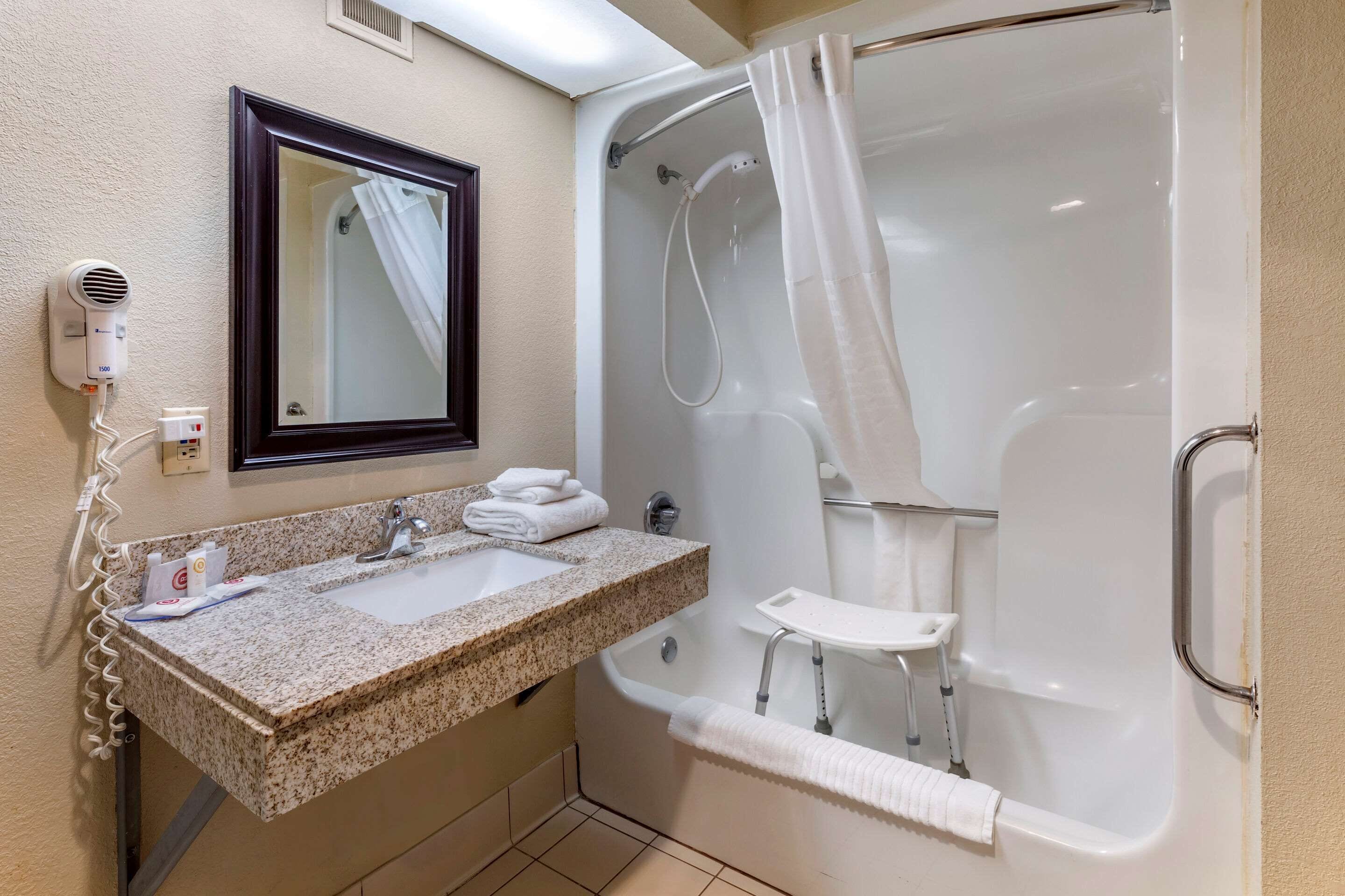 A bathroom with a granite sink, a mirror, a hair dryer, a shower with a glass door, and a small white stool.