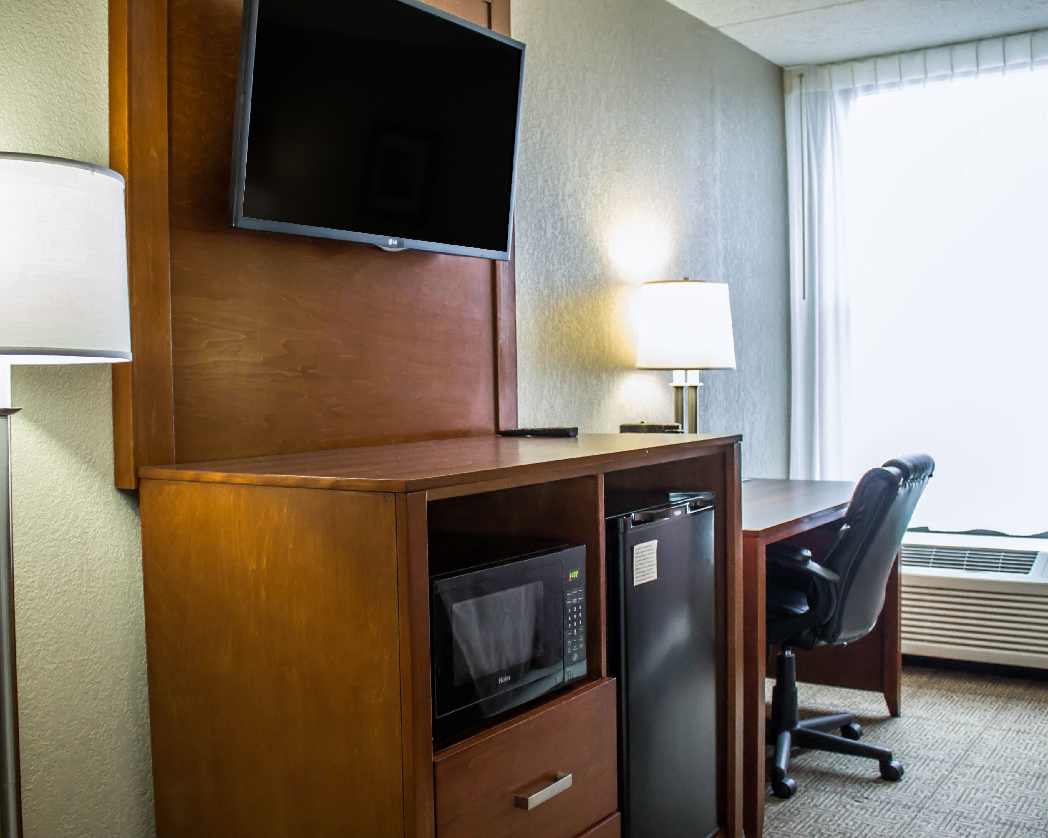 Une chambre d’hôtel avec un bureau en bois, un téléviseur fixé au-dessus, un four à micro-ondes, un réfrigérateur, une chaise et une fenêtre avec des rideaux.