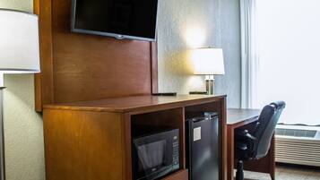 Une chambre d’hôtel avec un bureau en bois, un téléviseur fixé au-dessus, un four à micro-ondes, un réfrigérateur, une chaise et une fenêtre avec des rideaux.