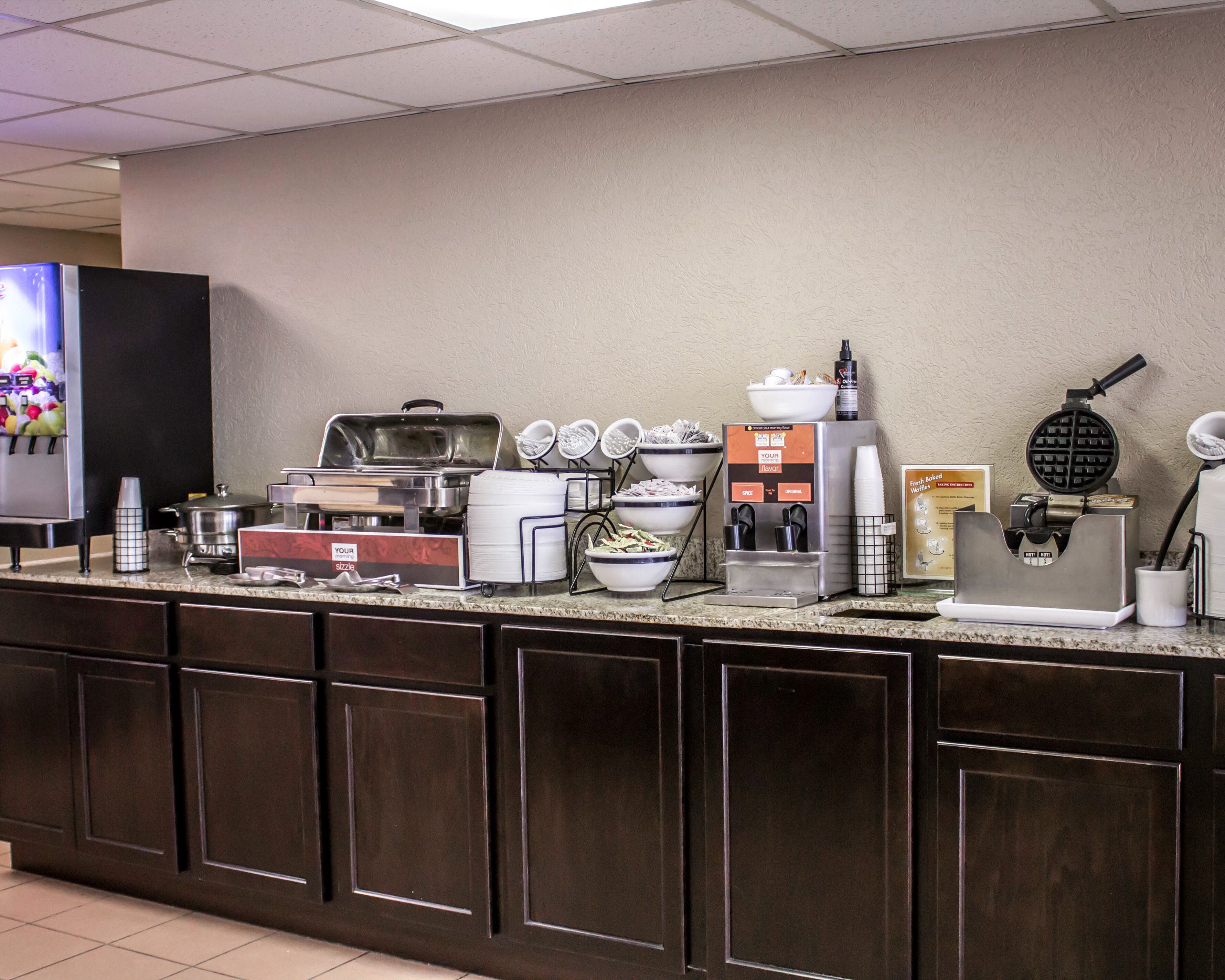 A self-service breakfast bar with various food and drink options, including a waffle maker, coffee machines, and bowls of cereal.