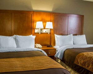 A hotel room with two beds, a wooden headboard, a nightstand with a phone and alarm clock, and two wall-mounted lamps.