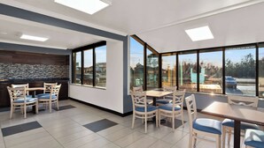 A modern dining area with wooden tables and chairs, large windows offering views of the outside, and a stone backsplash in the background. - Rodeway Inn Lumberton Central (Lumberton)