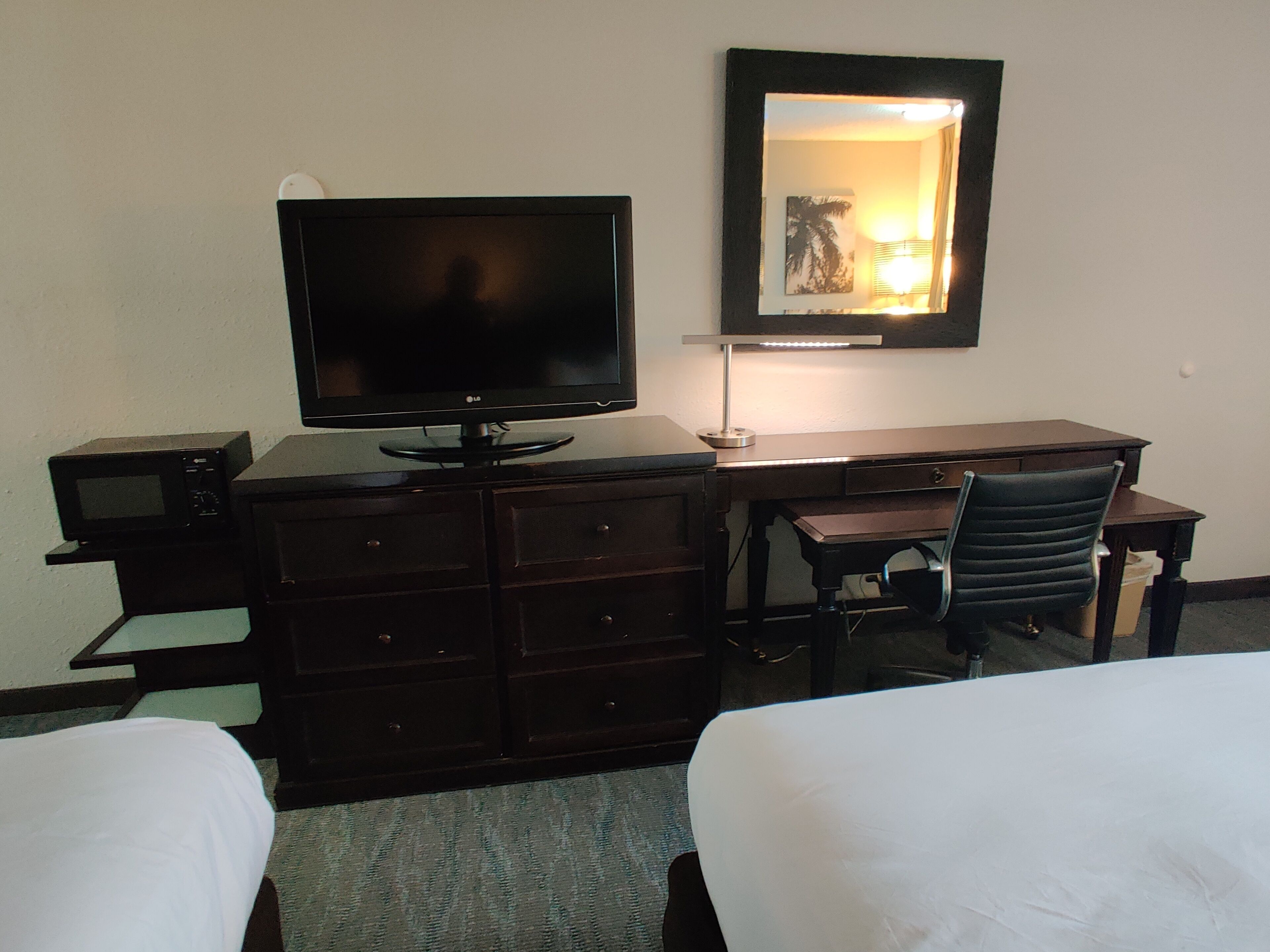Habitación de hotel con cama, escritorio con computadora y lámpara, televisión y espejo en la pared.