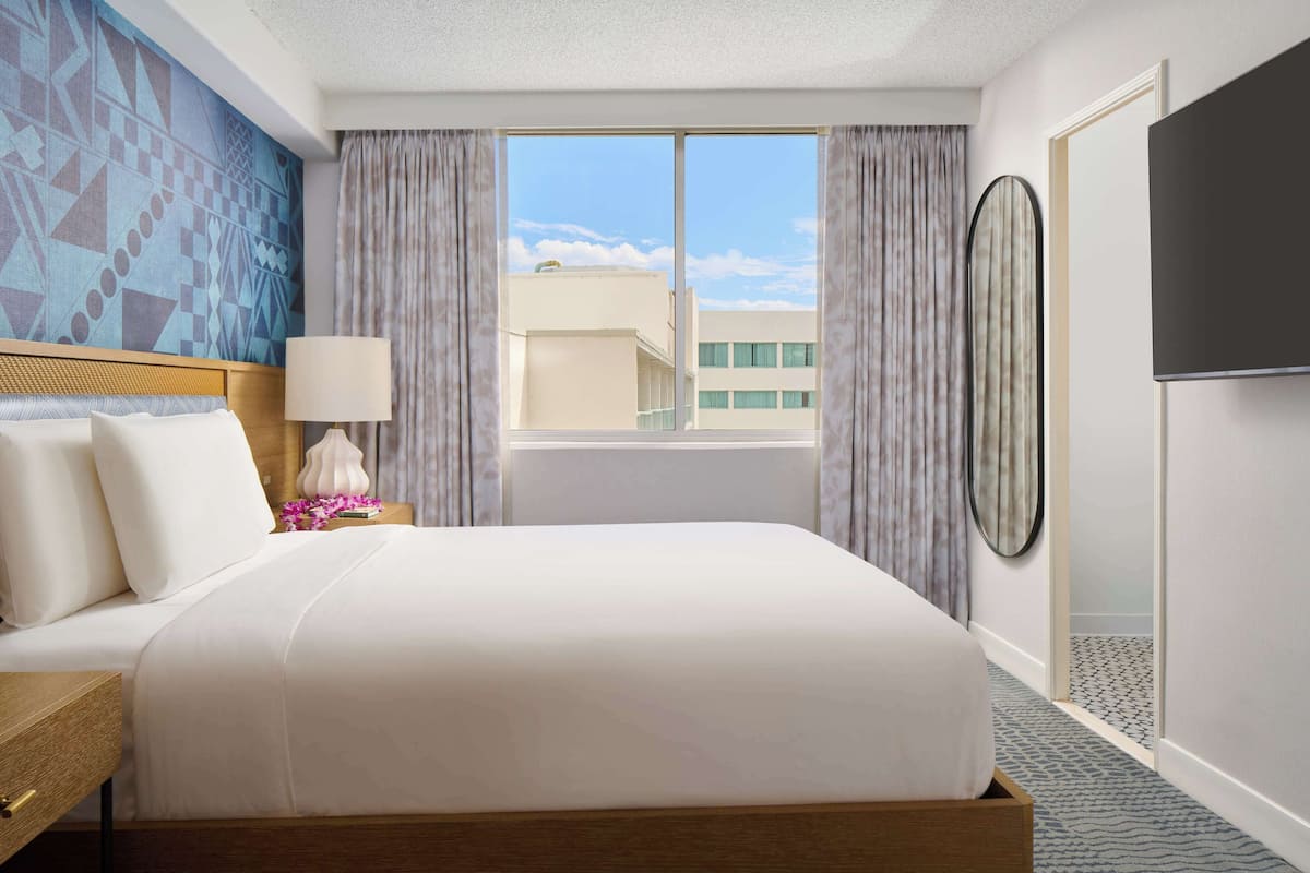 Une chambre d’hôtel avec un lit, une table de chevet, une lampe, un téléviseur, une fenêtre avec des rideaux et une vue sur des immeubles.
