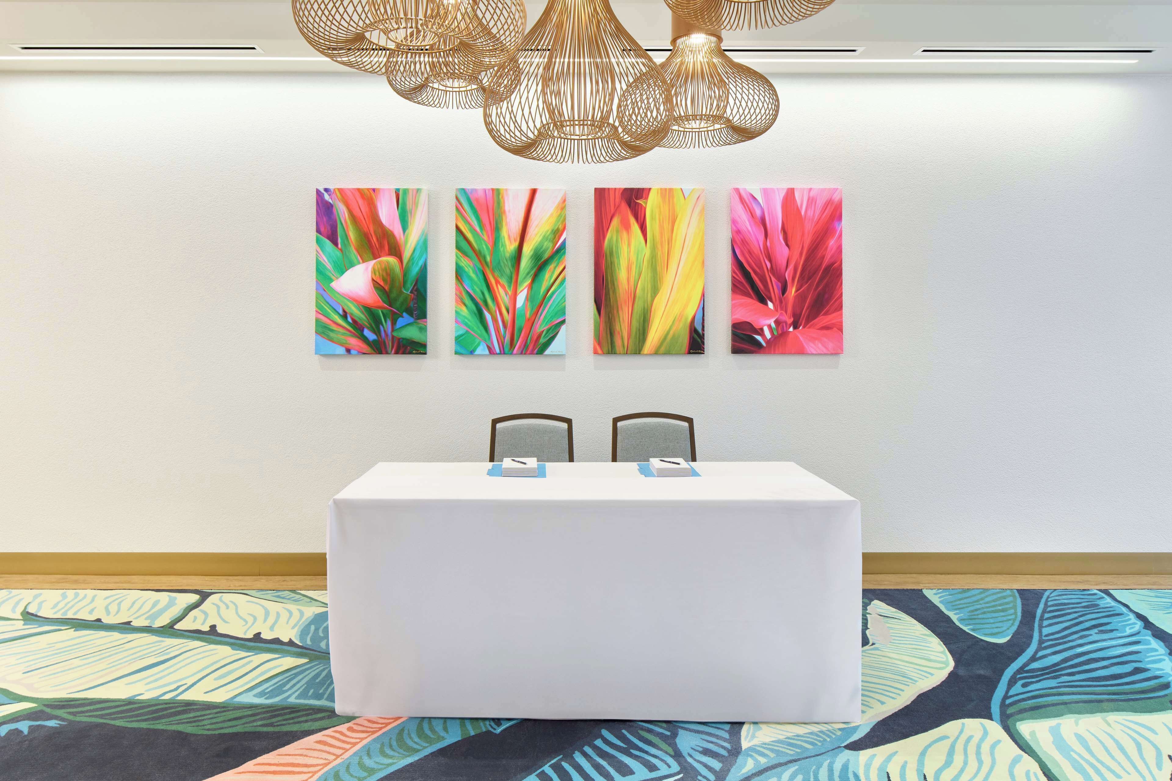 Une chambre d’hôtel avec une table blanche, deux chaises et un mur orné de trois tableaux colorés.