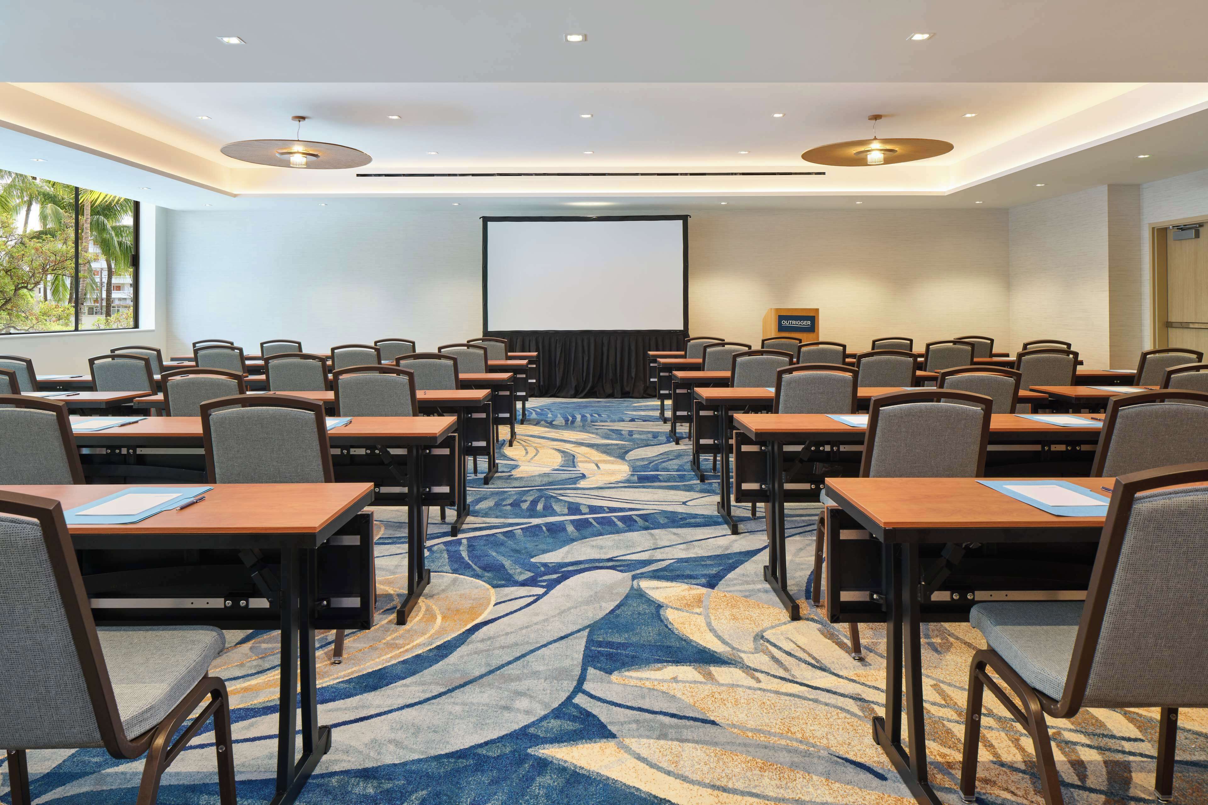 A conference room with rows of tables and chairs, a large screen, and a carpeted floor.