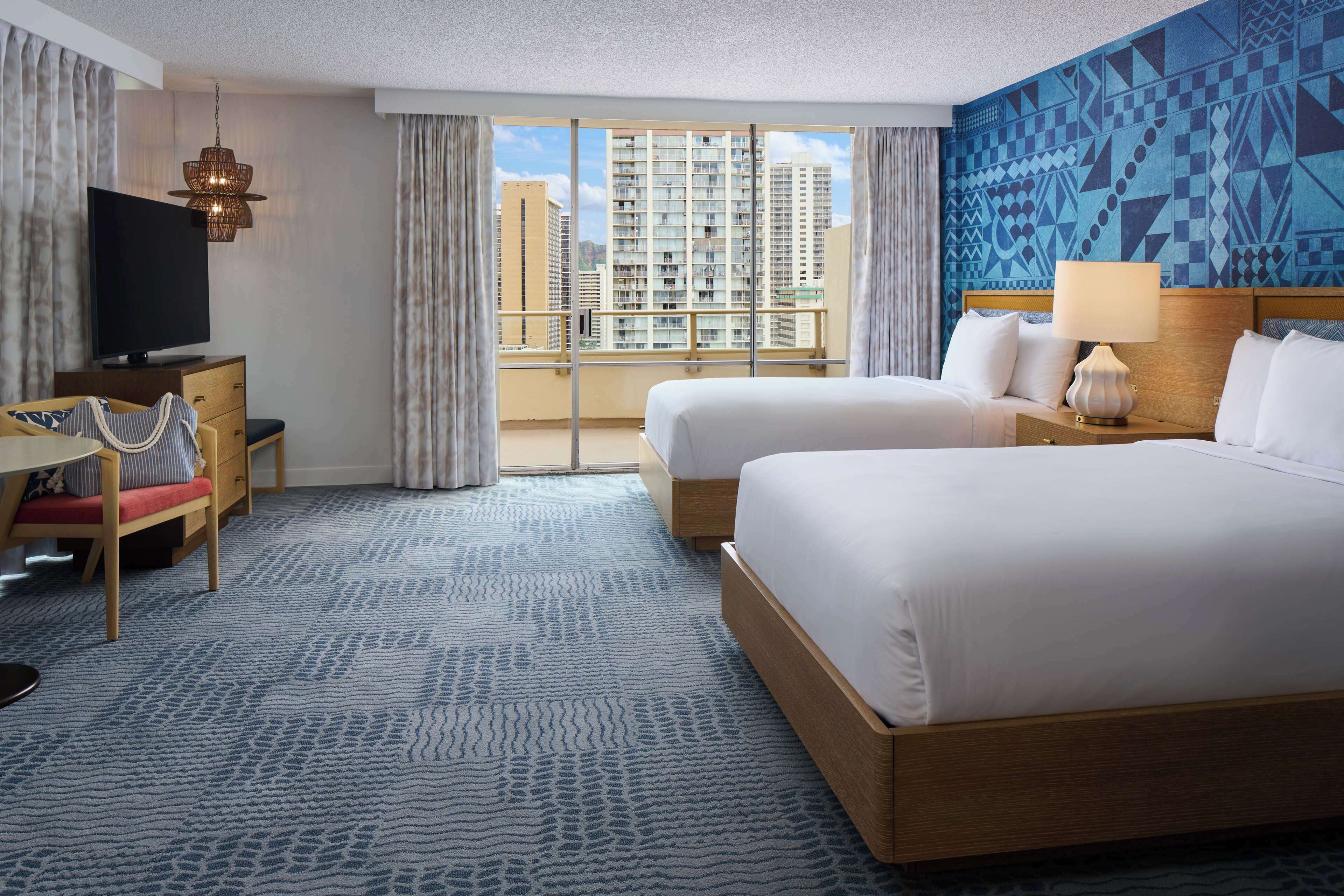 A hotel room with two beds, a TV, a desk, and a large window with a city view.