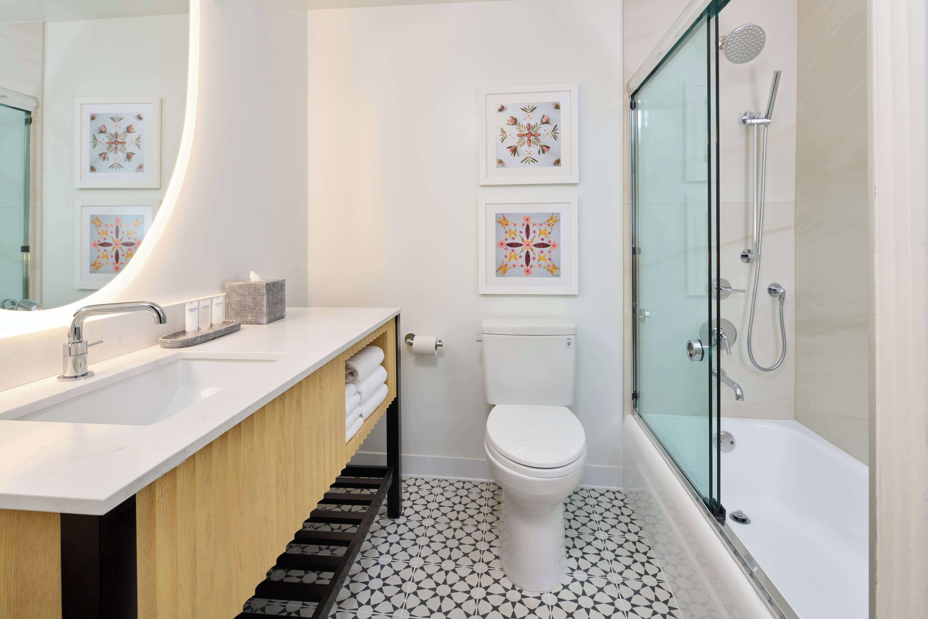 A modern bathroom with a white sink, toilet, and shower. There are framed artworks on the wall and a wooden vanity.
