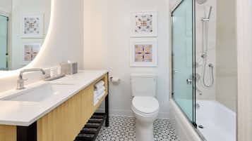 A modern bathroom with a white sink, toilet, and shower. There are framed artworks on the wall and a wooden vanity.