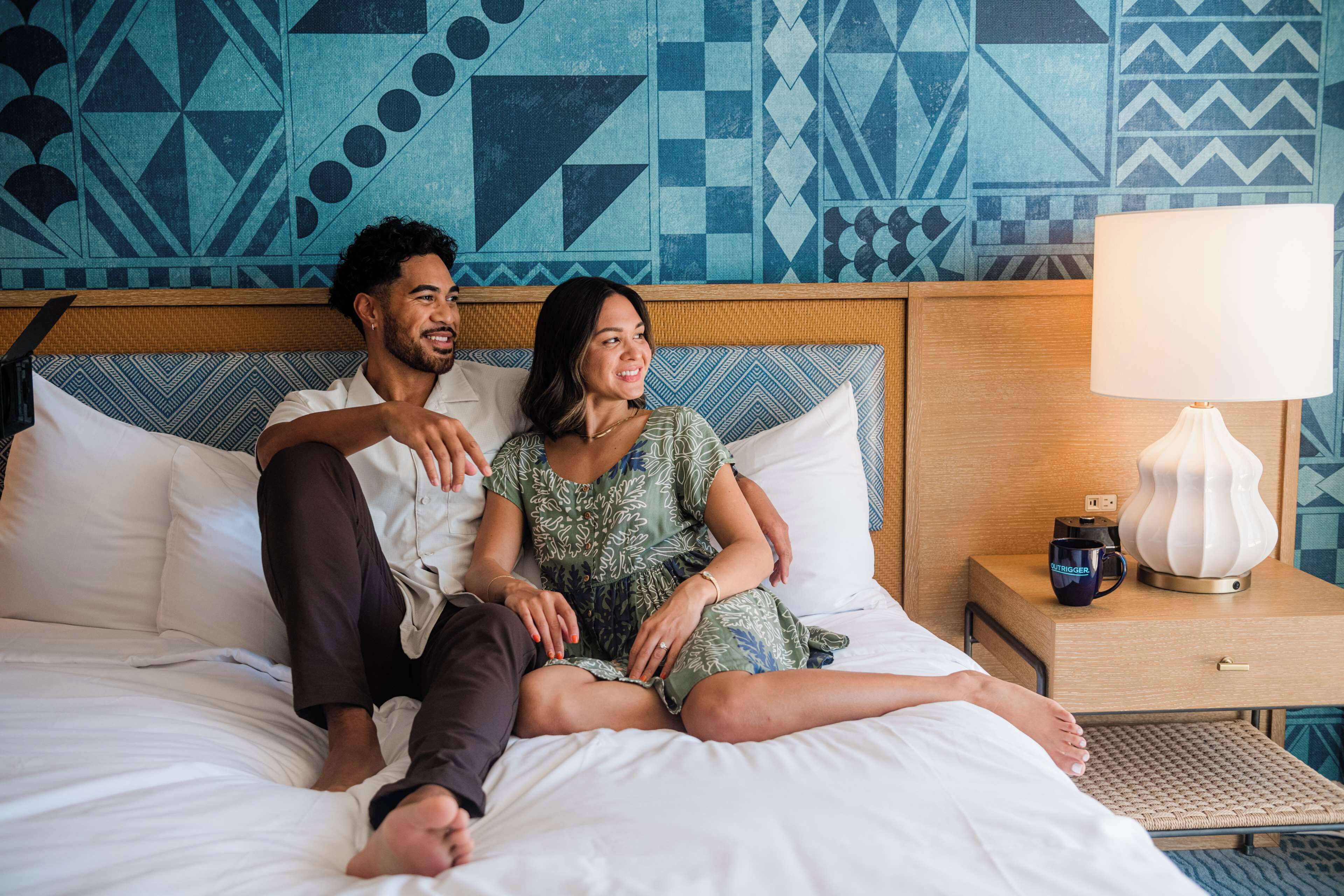 A bed with white bedding, a wooden headboard, a nightstand with a lamp, and a patterned wall.