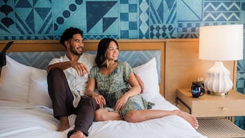 A bed with white bedding, a wooden headboard, a nightstand with a lamp, and a patterned wall.