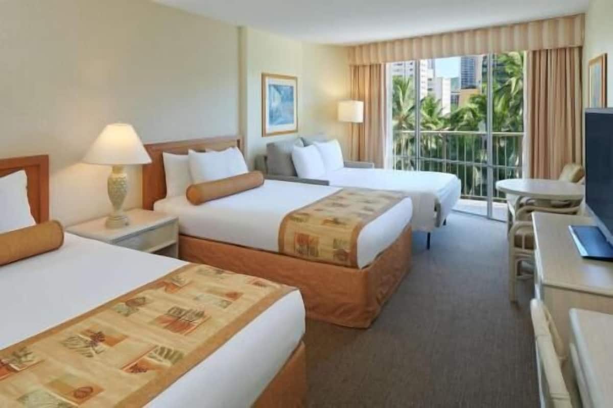 A hotel room with two beds, a desk, a chair, a TV, and a window with a view of buildings and greenery.