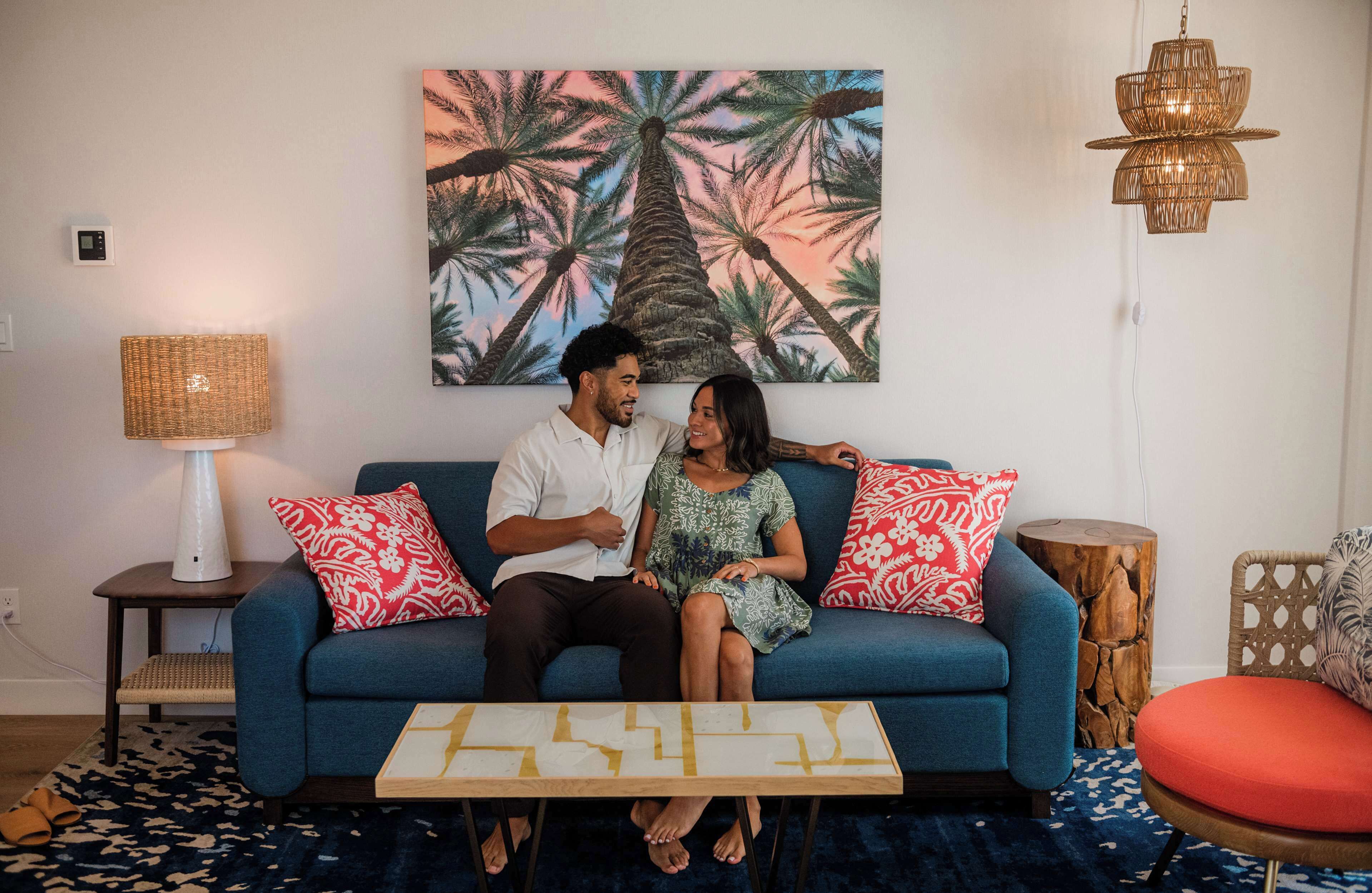 A couple sitting on a blue sofa with a tropical-themed painting above, a lamp, and a small table with a geometric design.