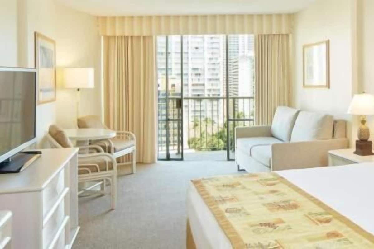 A hotel room with a bed, a desk with a computer, a chair, a sofa, and a balcony with curtains.
