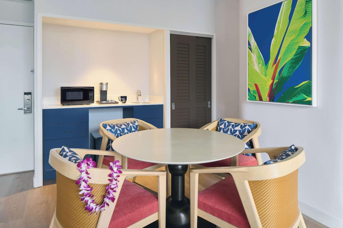 Une cuisine compacte équipée d’un four à micro-ondes, d’une machine à café et d’un petit comptoir. On y trouve une table ronde avec quatre chaises, chacune agrémentée d’un coussin à motifs. Une porte à persiennes est visible au fond.