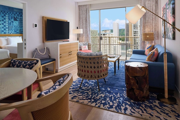 A modern hotel room with a large window, a sofa, a chair, a dining table, and a TV.