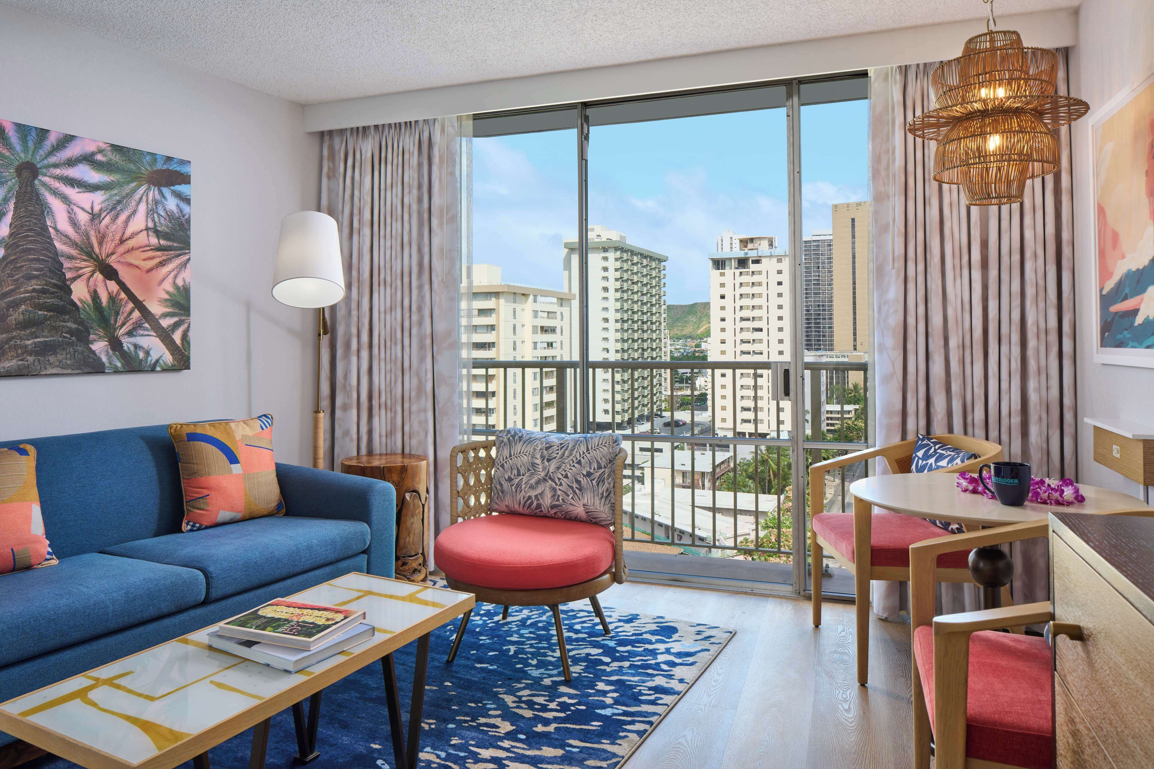 A modern hotel room with a balcony, a sofa, a chair, a table, and a large window offering a city view.