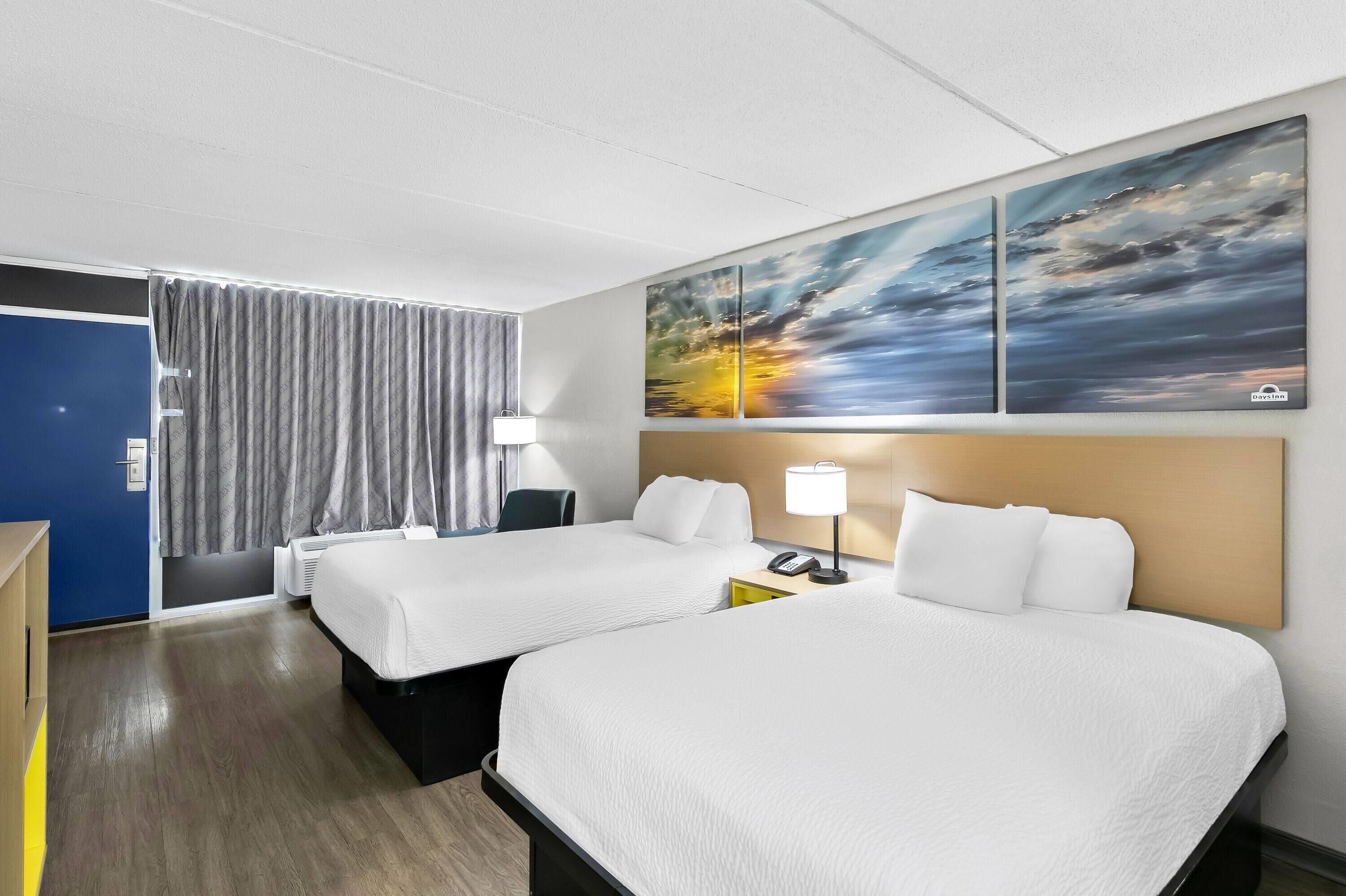 Quarto de hotel com duas camas, uma escrivaninha, um abajur e um quadro na parede.