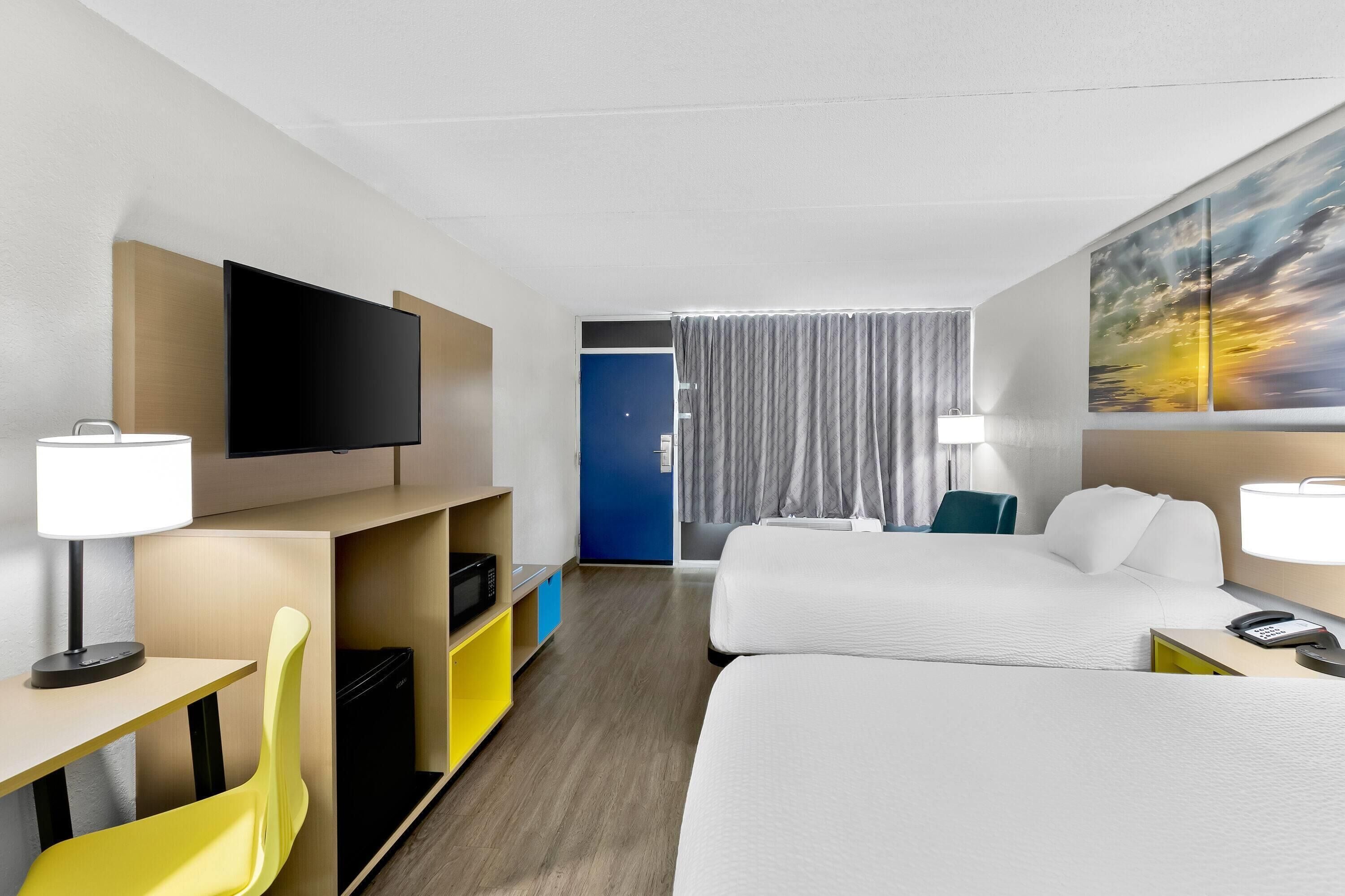 Habitación de hotel con dos camas, televisión, escritorio con lámpara, una silla y un cuadro en la pared.