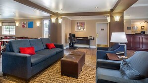 A hotel lobby with a dark blue sofa, a brown ottoman, and a grey armchair. There is a desk with a lamp, a chair, and a small table with a phone. The room has wooden flooring and a patterned rug. - Comfort Inn Castro Valley (Castro Valley)