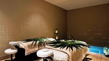 A spa room with a massage table, two footrests, a small table with a lamp, and a view of a swimming pool.