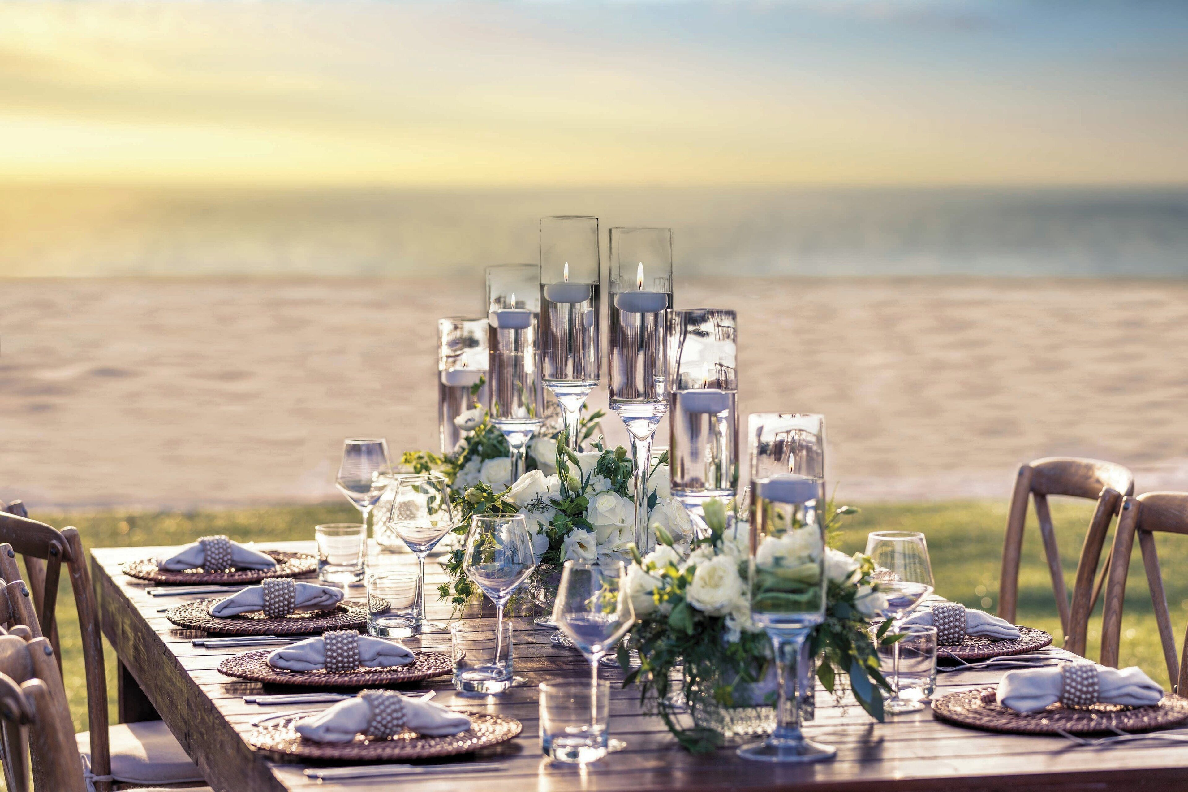 A table set for a meal with glasses, plates, and a centerpiece, overlooking a beach.
