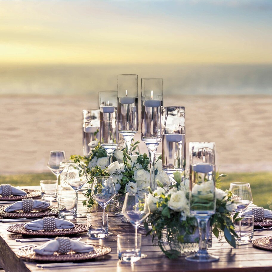A table set for a meal with glasses, plates, and a centerpiece, overlooking a beach.