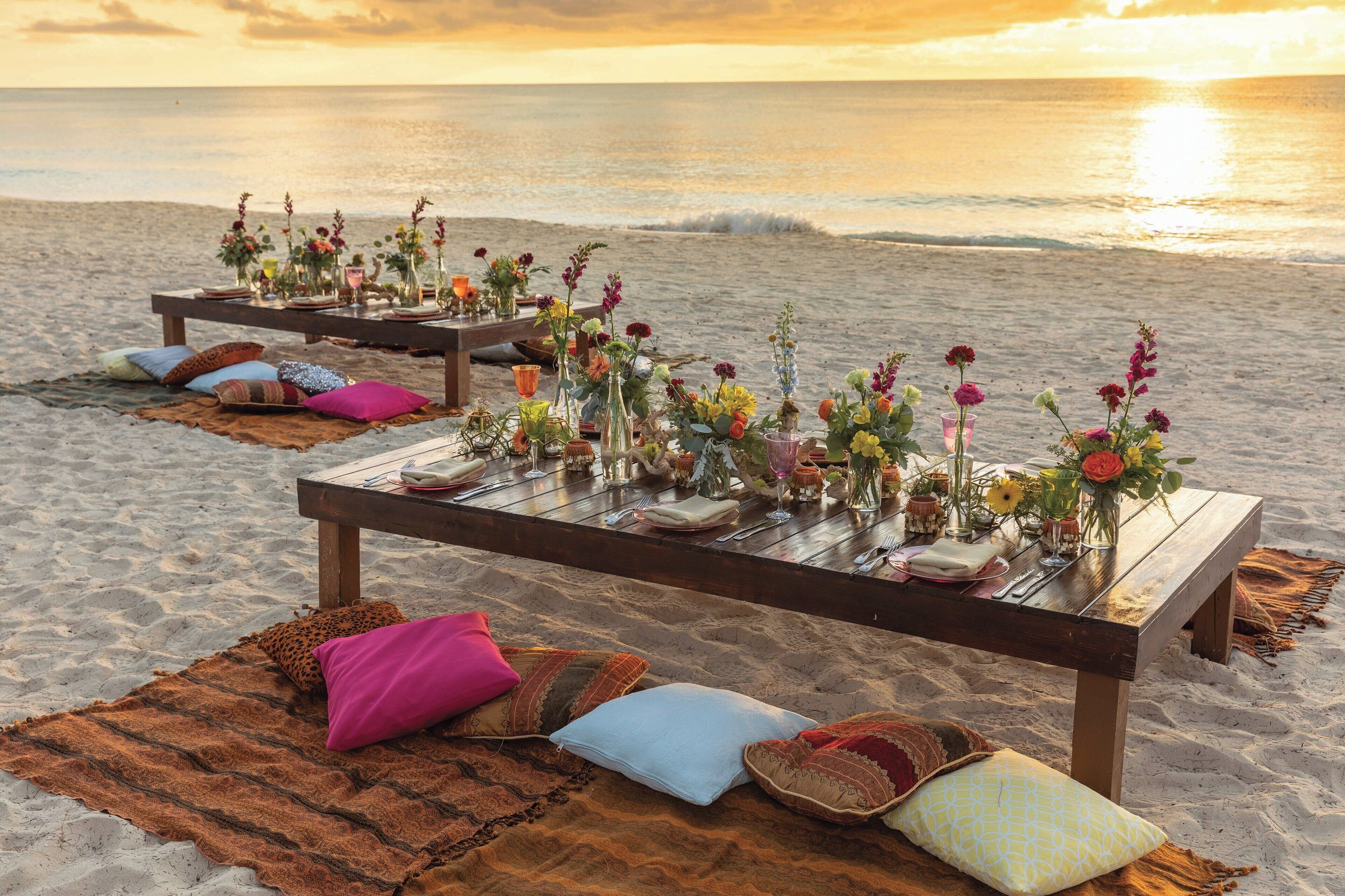 A beachside dining setup with wooden tables, colorful cushions, and floral arrangements.