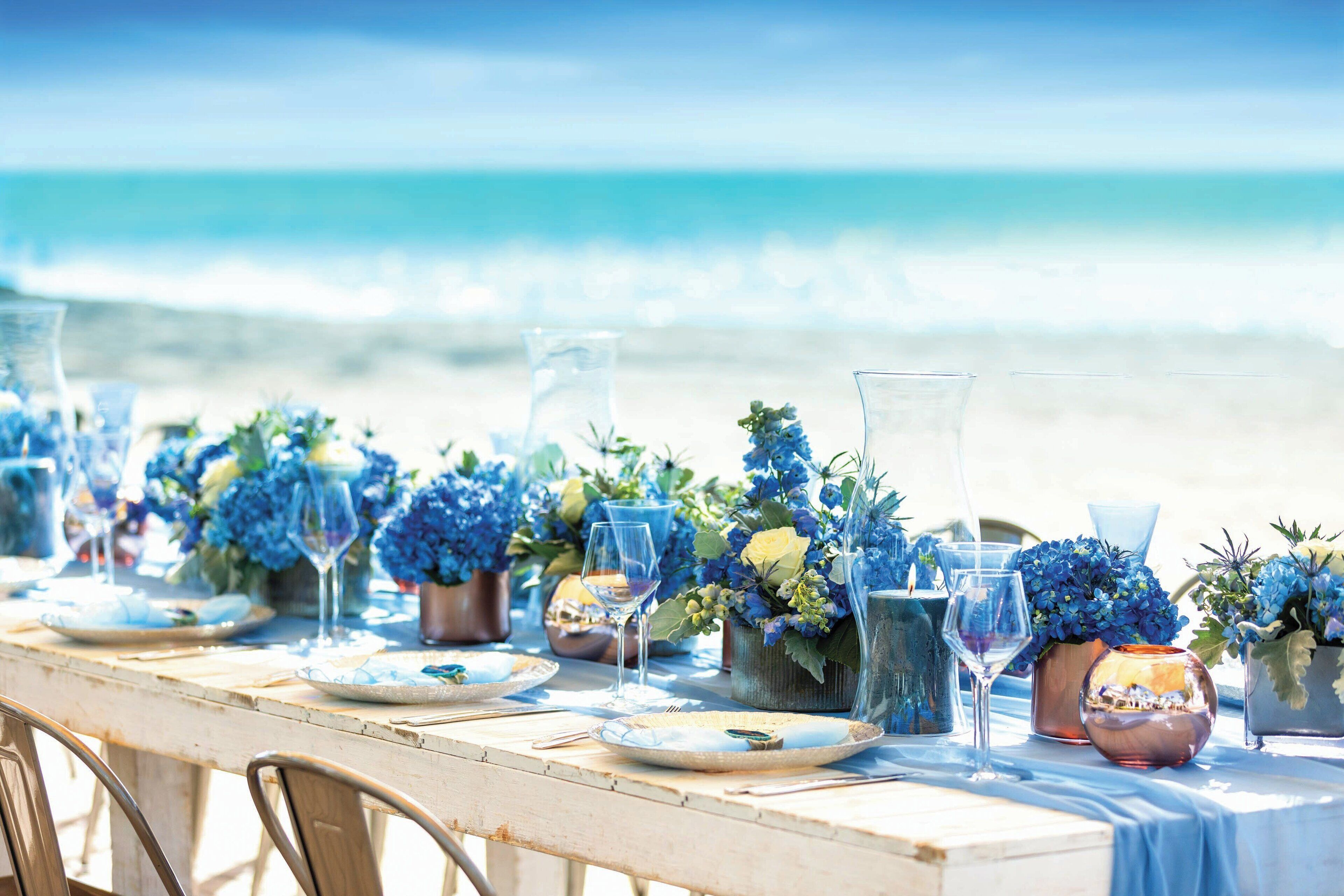 A beachside table set with blue flowers, clear glasses, and plates, overlooking the ocean.