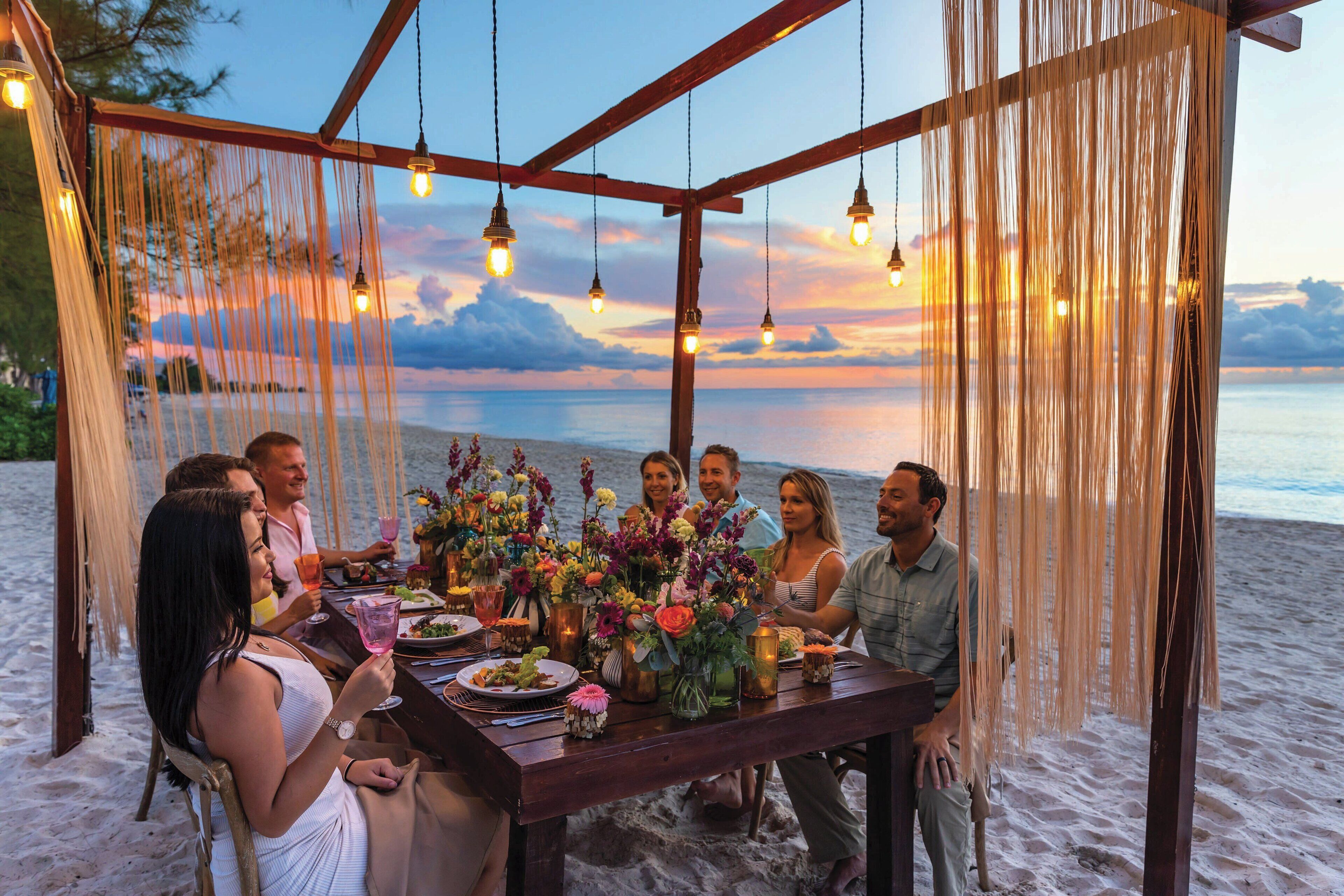 A group of people dining at a wooden table with hanging lights, set against a beach backdrop.