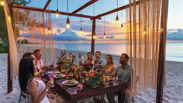 A group of people dining at a wooden table with hanging lights, set against a beach backdrop.